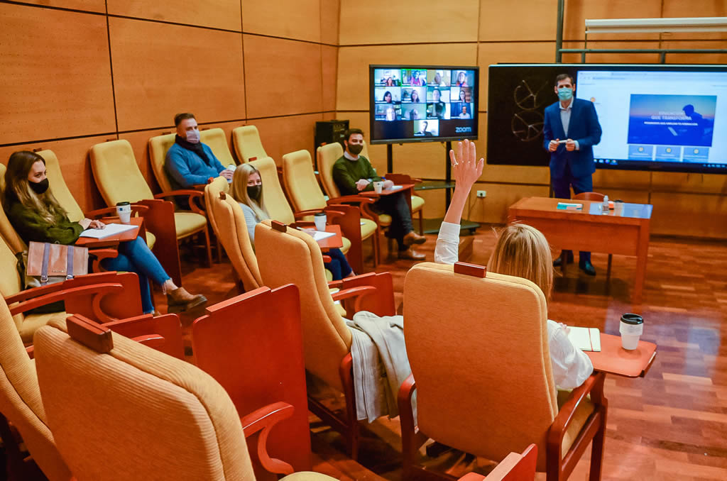 Un auditorio de la Escuela de Graduados con el profesor José Arnoletto vistiendo traje, de pie, y hay cuatro estudiantes sentados en los sillones, una de ellas levanta la mano y hay una pantalla con una sesión de Zoom con una docena de estudiantes conectados y una pantalla paralela con contenidos de la clase
