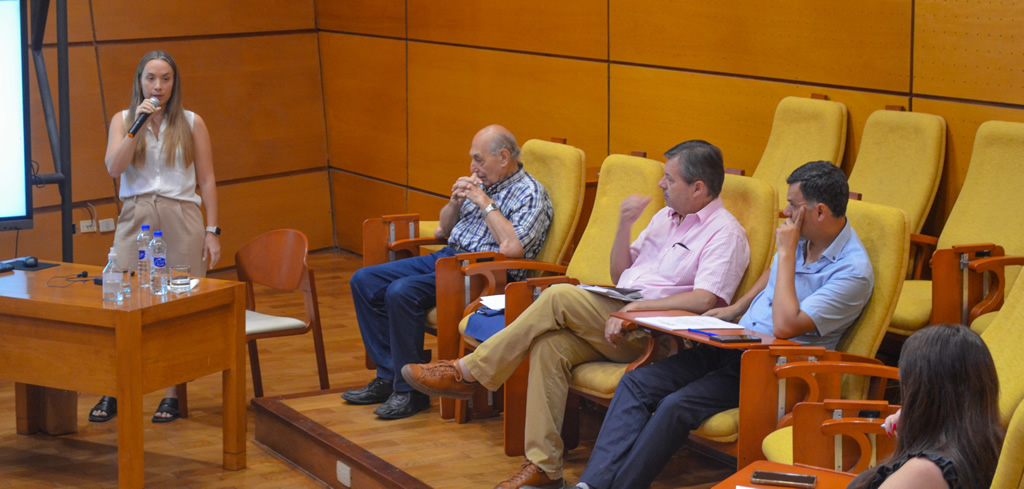 Virginia Giordano usando un micrófono hablando de pie en un auditorio de la Escuela de Graduados con paredes y pisos de madera, y delante de ella hay una mesa con tres botellas de agua mineral mientras que sentados la escuchan tres profesores y una profesora