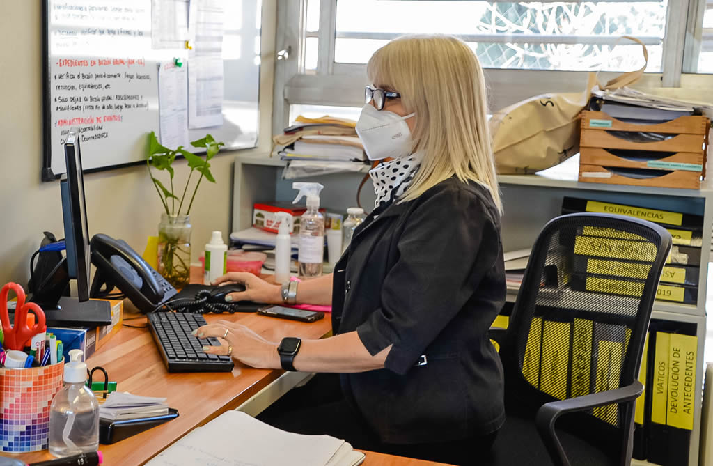 Una trabajadora nodocente sentada con barbijo y anteojos en un día soleada tipeando sobre un teclado de computadora frente a un monitor y hay una pizarra en la pared más un armario con carpetas de archivos