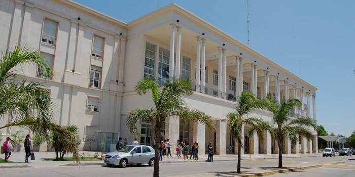 Edificio del Pabellón Argentina de Ciudad Universitaria en un día soleado con gente caminando en la vereda y automóviles circulando en la calle