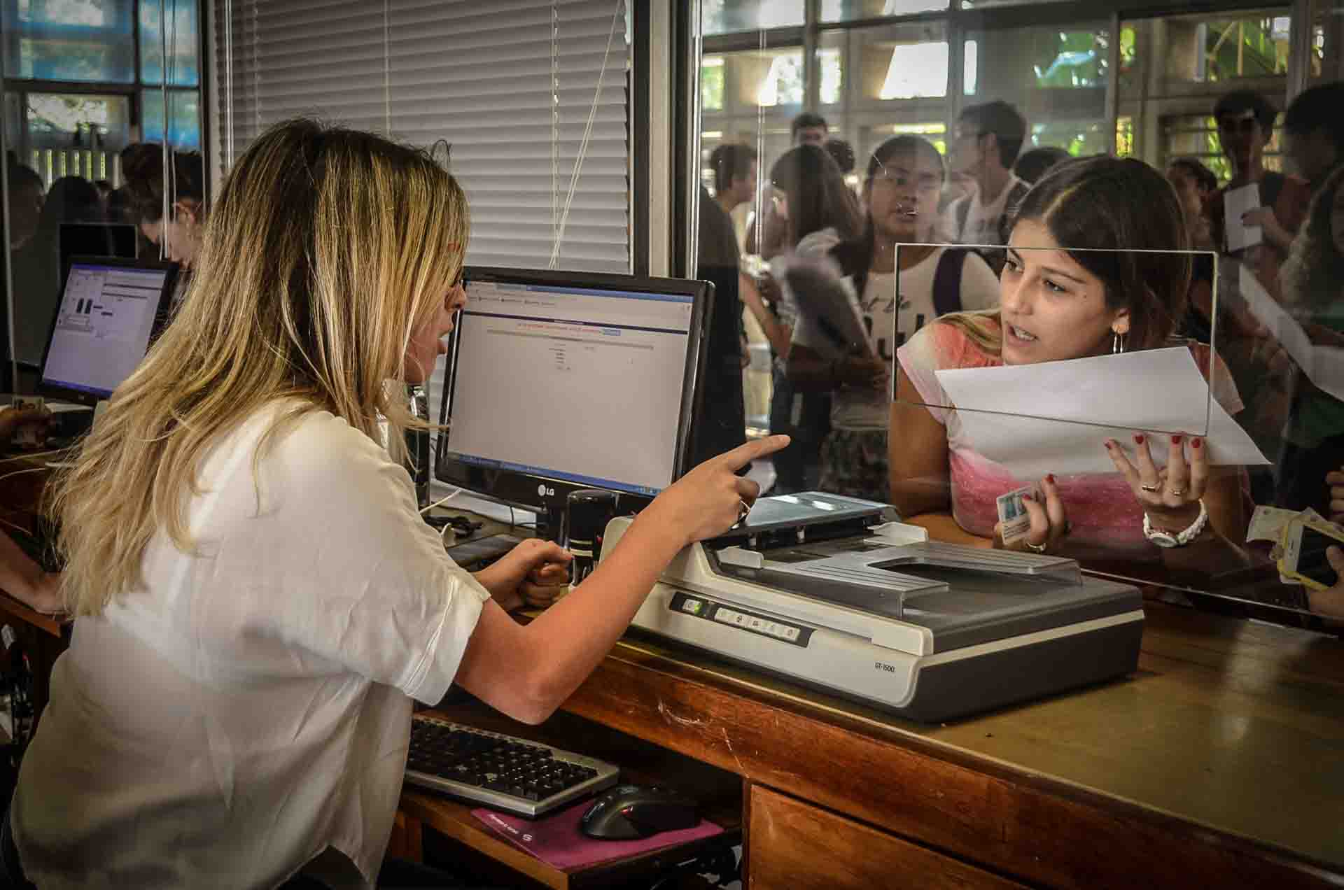 Una trabajadora nodocente atiende tras el vidrio a una estudiante que entrega documentación