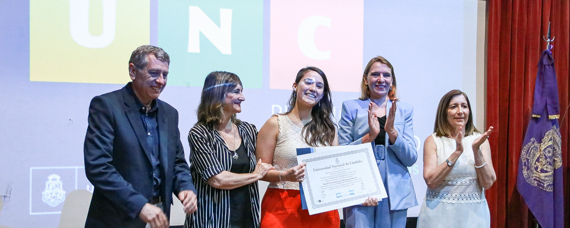 La Vicerrectora Marchisio junto a una egresada premiada y tres autoridades más aplaudiendo y detrás hay una pantalla gigante junto a una bandera con el escudo de la Universidad Nacional de Córdoba