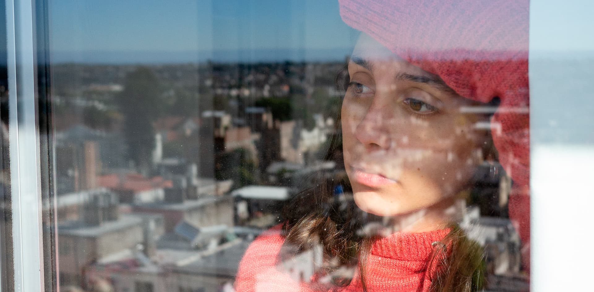 Una estudiante apenada con un gorro de lana y pulover mirando detrás de una ventana en un día soleado