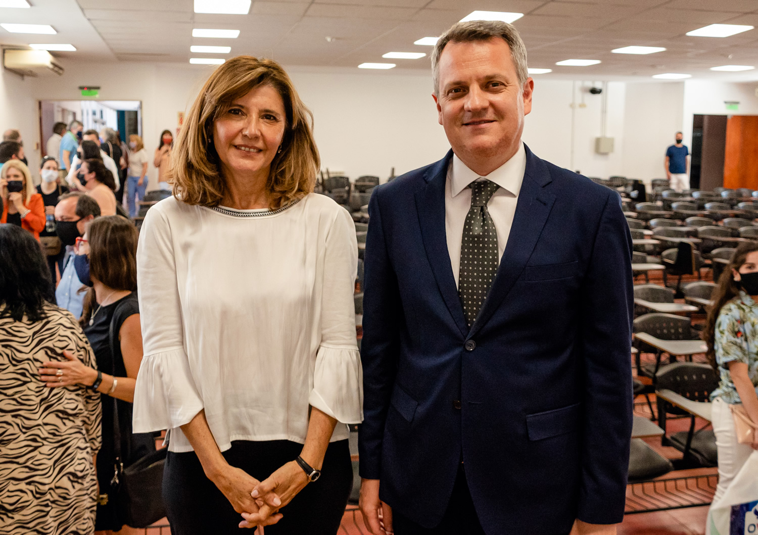 Ricardo Descalzi con saco y corbata y Catalina Alberto con blusa sonrientes de pie dentro de un aula con muchas butacas vacías y una veintena de personas de pie y caminando en un día soleado