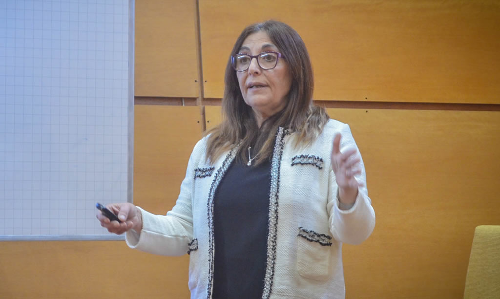 Inés Asís con lentes  y cabello largo vistiendo un saco de lana expone y explica un tema con un marcador en su mano derecha delante de una pizarra de acrílico y paneles de madera