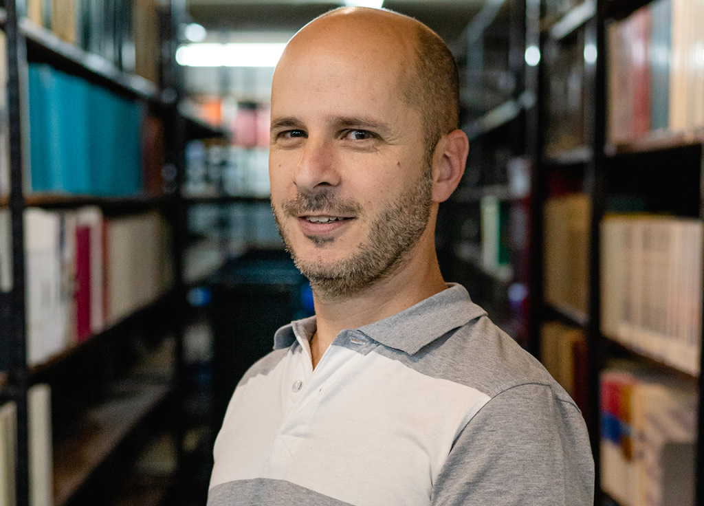 David Giuliodori sonriendo vistiendo una chomba y detrás hay dos estanterías con libros