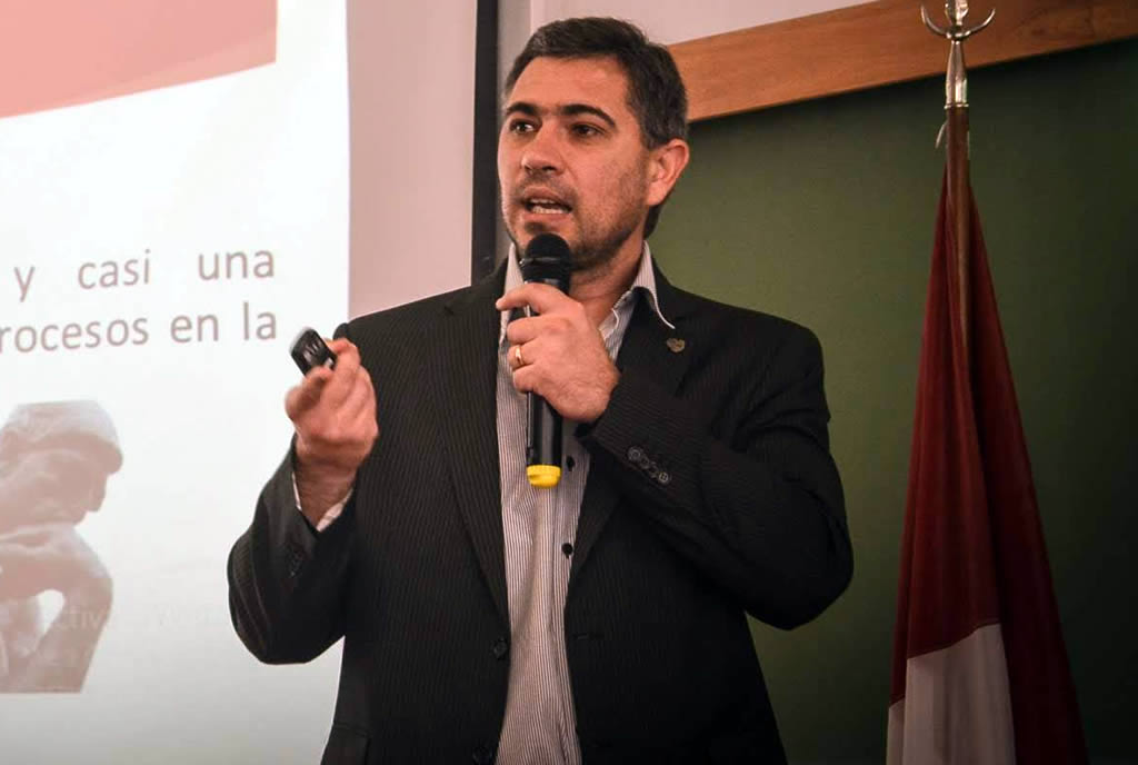 Ernesto Paiva exponiendo vistiendo un saco y camisa, sosteniendo un micrófono sin cable y un puntero láser, delante de un pizarrón y una bandera de la provincia de Córdoba en un mástil