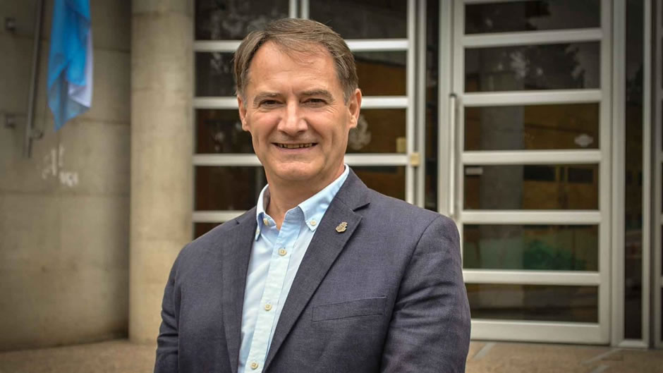 Gerardo Heckmann de pie, sonriente, vistiendo saco, en frente de la Escuela de Graduados, de día, y hay una bandera de Argentina detrás