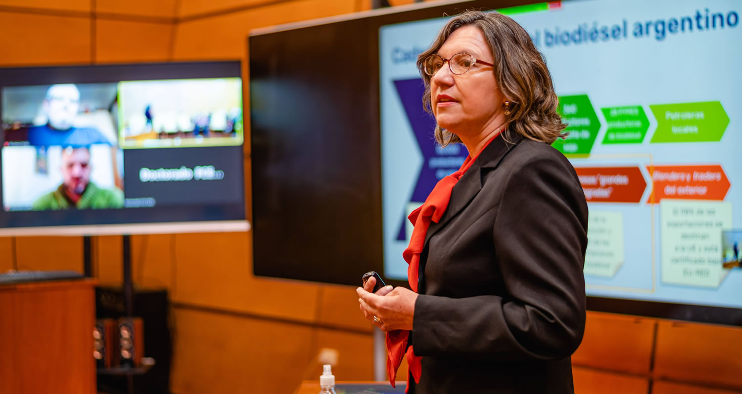 Mónica Buraschi presenta su tesis doctoral, usando anteojos, y detrás hay dos pantallas