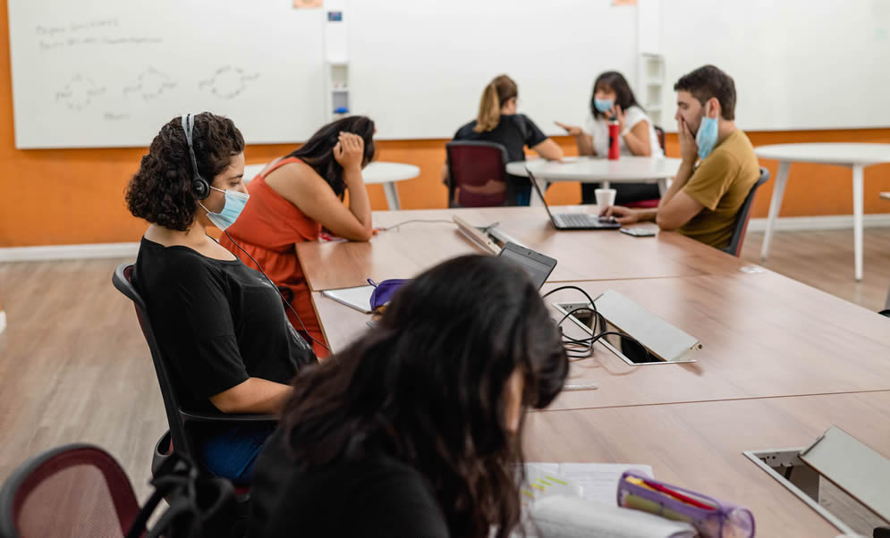 En una mesa grande de la Biblioteca hay cuatro estudiantes sentados con barbijos leyendo y escribiendo y al fondo en una mesa redonda hay dos estudiantes mujeres con barbijos conversando