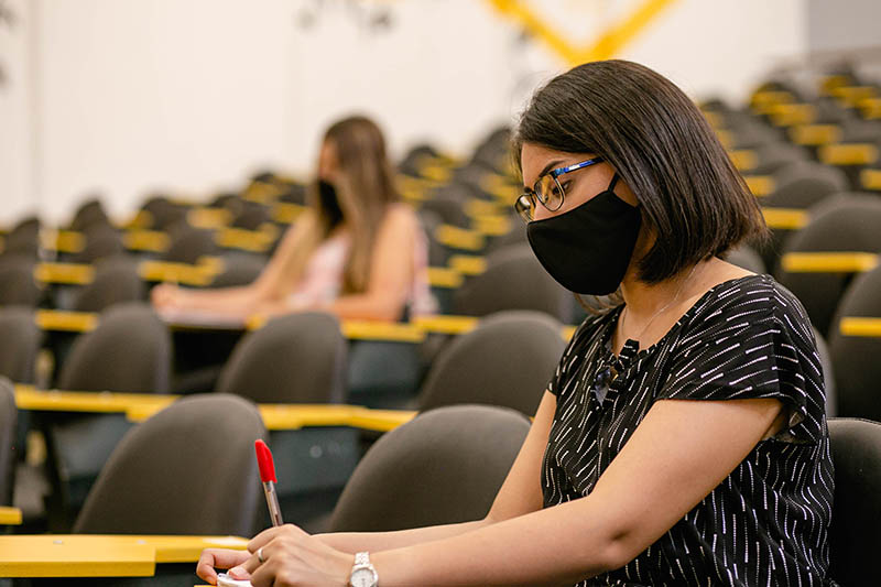 Una estudiante con barbijo en una de las aulas renovadas de la Facultad anota sentada en un pupitre y cinco metros detrás hay otra estudiante tomando apuntes