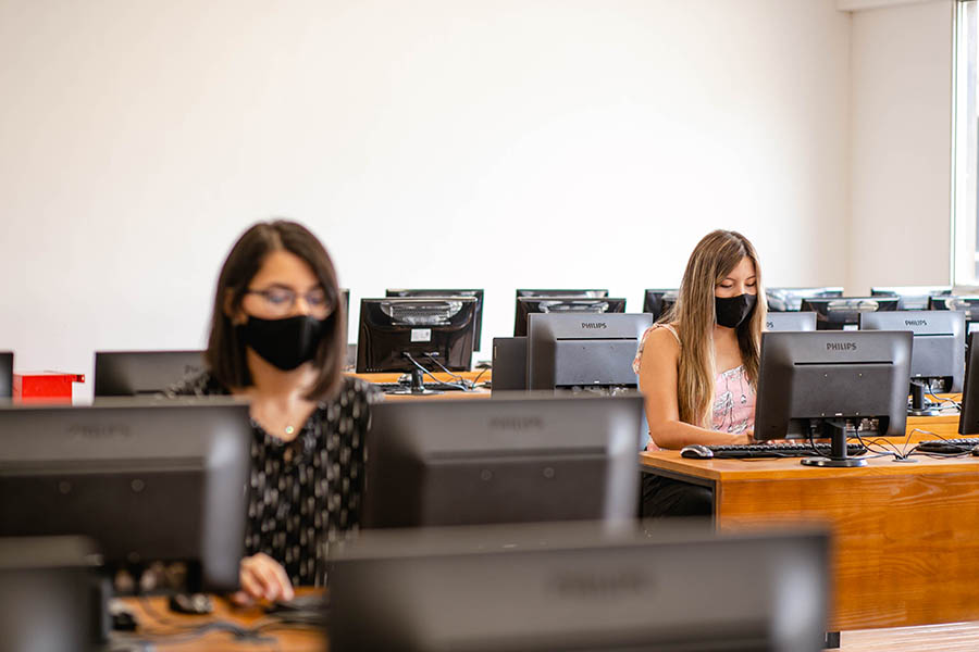 Dos estudiantes mujeres con barbijos sentadas usando computadoras en un día luminoso