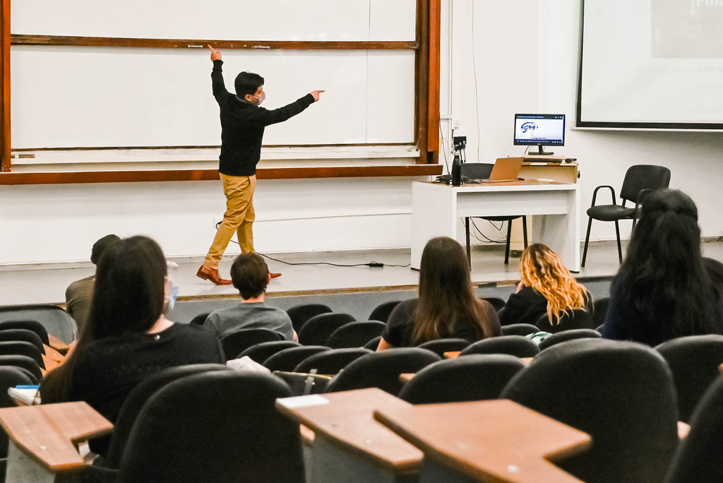 Un docente de pie señalando un pizarrón ante la mirada de una decena de estudiantes sentados con barbijo y hay un escritorio que tiene un monitor