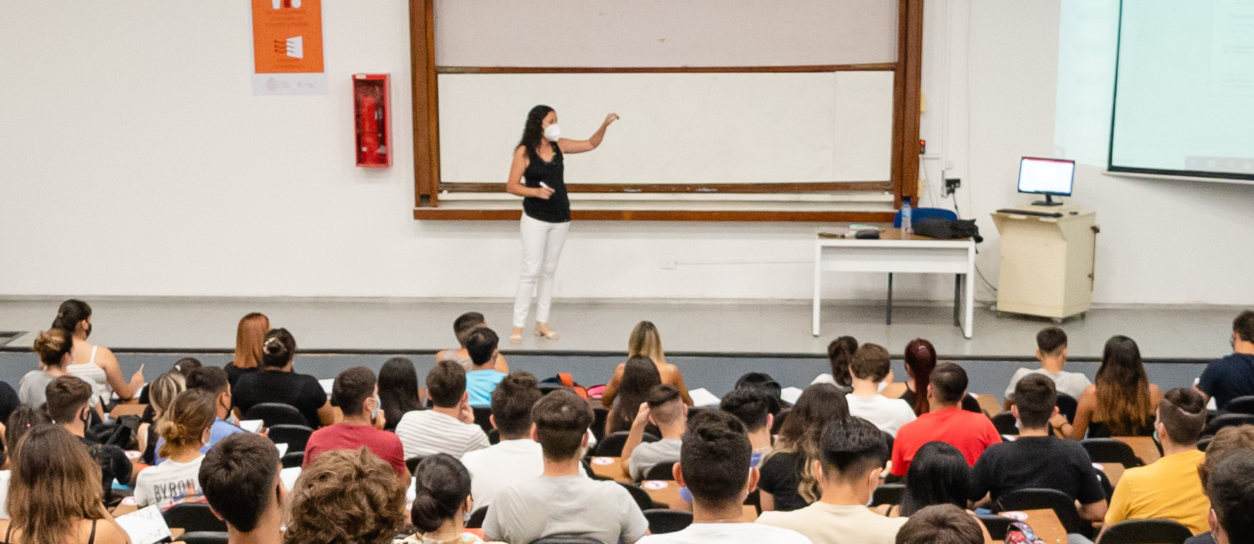 Una docente con barbijo de pie en un aula señala una pantalla gigante ante la mirada de una treintena de estudiantes sentados