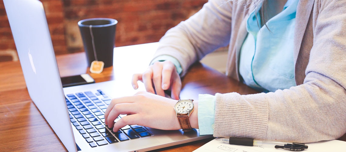 Una mujer tipeando en una computadora portátil con una taza de té sobre el escritorio